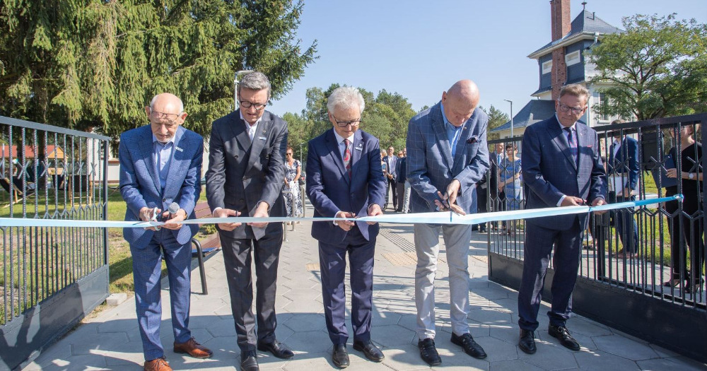 Pięciu mężczyzn przecina symboliczną wstęgę rozciągniętą przez bramę wjazdową na teren Centrum. Przecięcie wstęgi symbolizuje otwarcie Centrum.