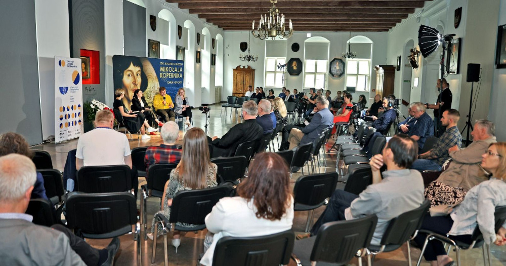 Wnętrze Sali Wielkiej w Ratuszu Staromiejskim w Toruniu, w której odbywa się debata trzech mężczyzn i jednej kobiety. Wokół debatujących siedzą obserwujący uczestnicy.