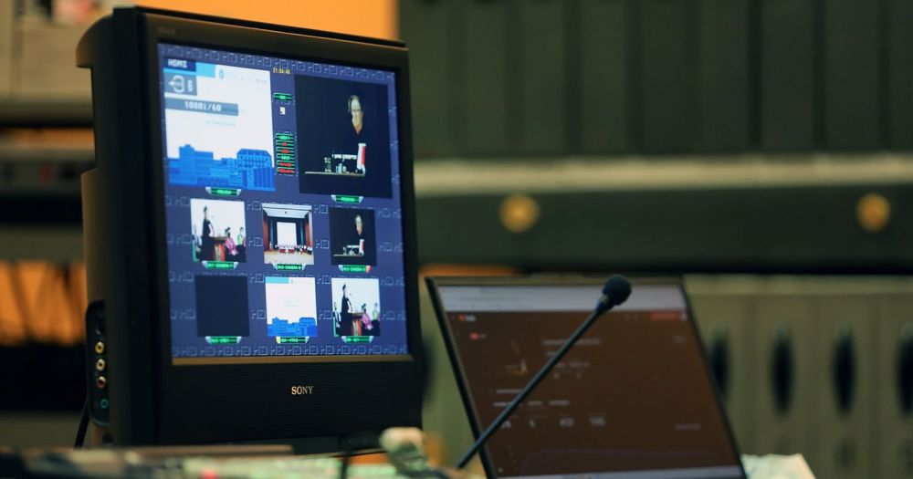 Na zdjęciu widoczny jest ekran z nagrywanym wystąpieniem, mikrofon, ekran laptopa