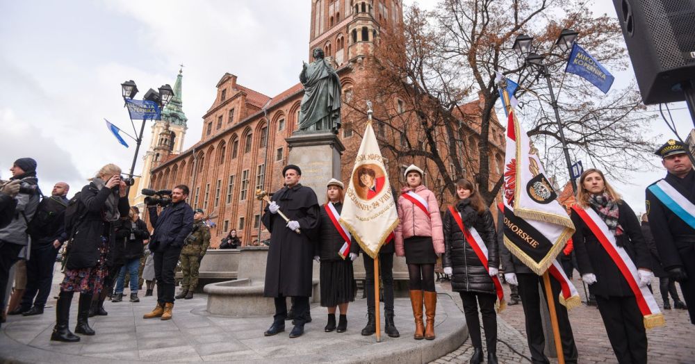 Osoby trzymające sztandar stoją pod pomnikiem Mikołaja Kopernika w Toruniu