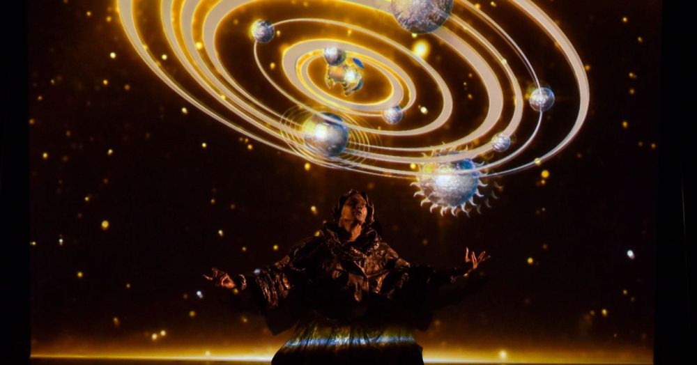 A man dressed as Nicolaus Copernicus on stage. A visualization of the Solar System is displayed above it.