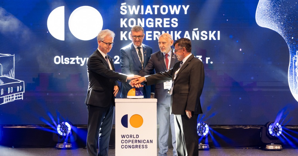 Czterech mężczyzn stoi na scenie i trzyma wyciągnięte dłonie nad specjalnym przyciskiem nawiązującym do logotypu Światowego Kongresu Kopernikańskiego. Jego wciśnięcie ma być symbolem rozpoczęcia Kongresu.