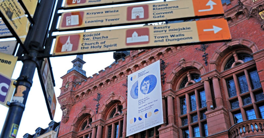 Na pierwszym planie drogowskazy wskazujące drogę do różnych instytucji kultury w mieście. Na drugim planie fasada Dworu Artusa w Toruniu z banerem Światowego Kongresu Kopernikańskiego.